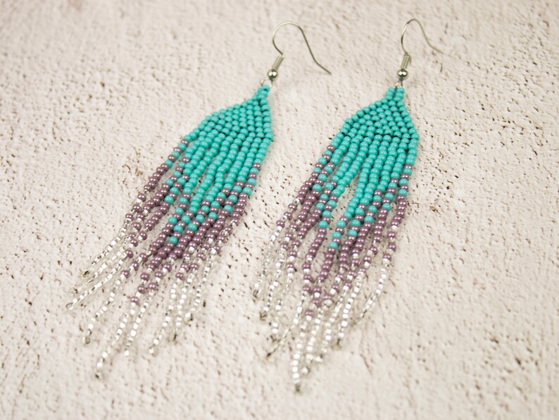 May include: A pair of long, beaded earrings with a gradient of turquoise, purple, and silver beads. The earrings are made with small, round beads and have a silver hook fastening.