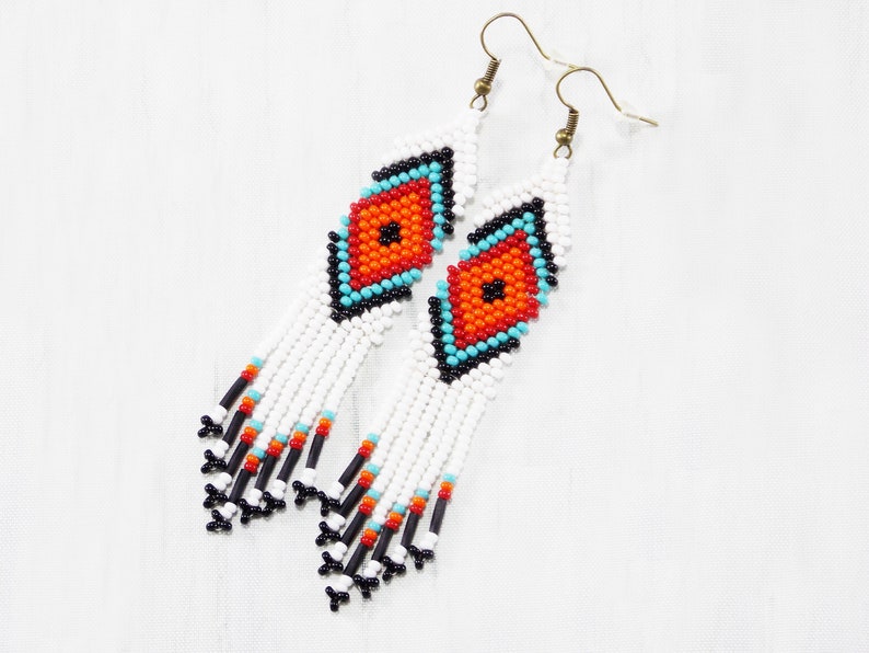 May include: Handmade beaded earrings with a diamond pattern. The earrings feature a white base with a central diamond design in orange, turquoise, and black. The fringe ends with black and orange beads.