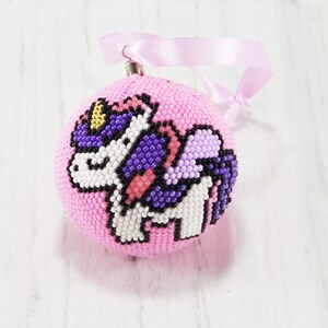 May include: A pink beaded Christmas ornament with a unicorn design. The unicorn is white with purple and pink wings and a gold horn.