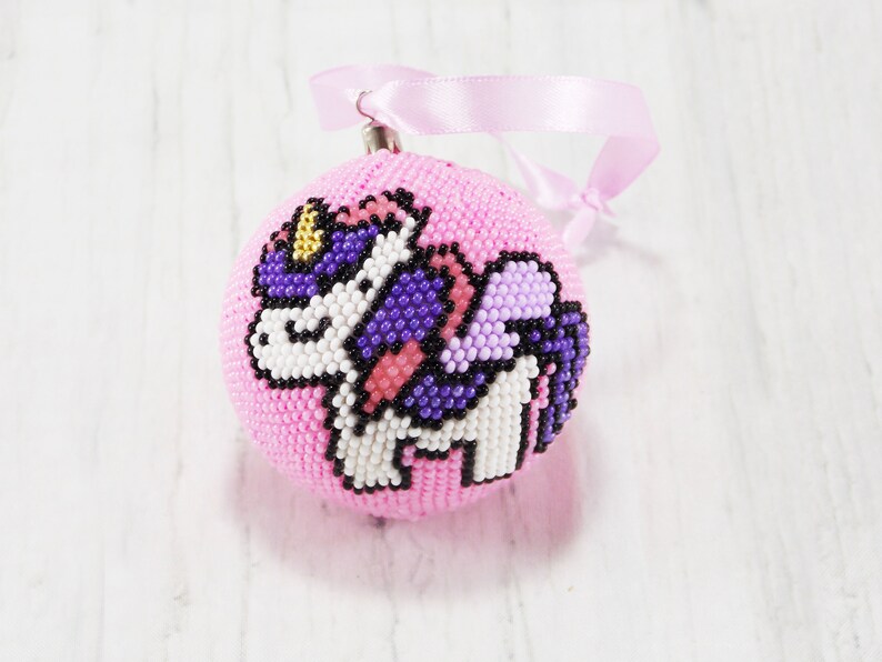 May include: A pink beaded Christmas ornament with a unicorn design. The unicorn is white with purple and pink wings and a gold horn.