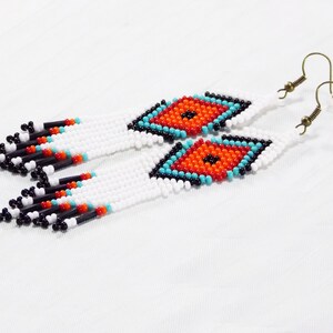 May include: White beaded earrings with a diamond-shaped design in orange, turquoise, and black. The earrings have a fringe of black, red, and white beads. The ear hooks are a bronze color.