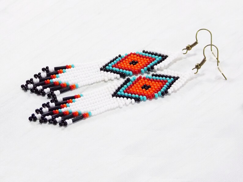 May include: White beaded earrings with a diamond-shaped design in orange, turquoise, and black. The earrings have a fringe of black, red, and white beads. The ear hooks are a bronze color.
