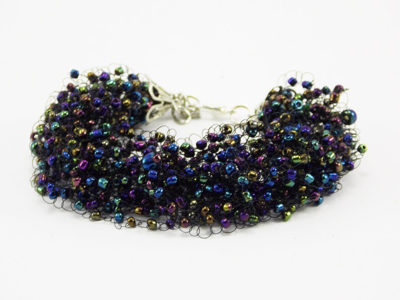 May include: A black beaded bracelet with iridescent beads in shades of blue, green, and purple. The bracelet is made with a stretchy cord and has a silver clasp.