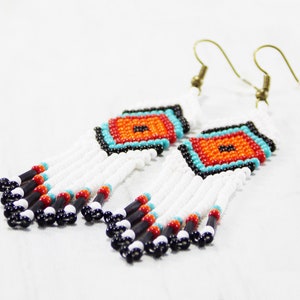 May include: Handmade beaded earrings with a geometric design. The earrings feature a central square with orange, turquoise, and black beads. White beads form the fringe, with black, red, and turquoise accents. The earrings have gold-colored ear wires.