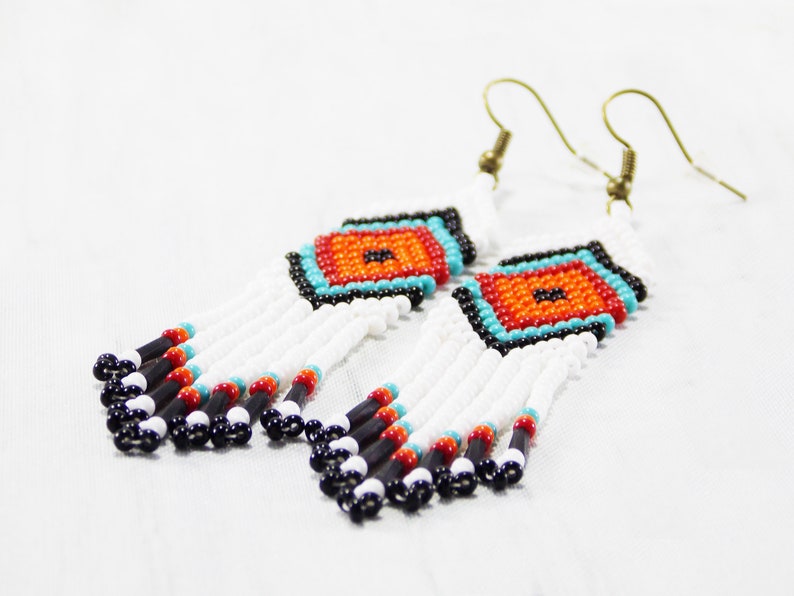 May include: Handmade beaded earrings with a geometric design. The earrings feature a central square with orange, turquoise, and black beads. White beads form the fringe, with black, red, and turquoise accents. The earrings have gold-colored ear wires.