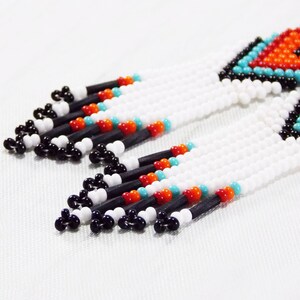 May include: Close-up of beaded earrings with a geometric design. The earrings feature white, black, orange, and turquoise beads. The top section has a diamond pattern in orange and turquoise. The fringe is white with black, orange, and turquoise accents.