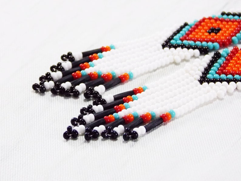 May include: Close-up of beaded earrings with a geometric design. The earrings feature white, black, orange, and turquoise beads. The top section has a diamond pattern in orange and turquoise. The fringe is white with black, orange, and turquoise accents.