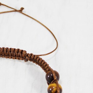May include: A brown braided cord bracelet with tiger's eye beads. The bracelet is tied with a slip knot.