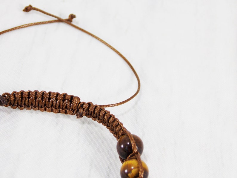 May include: A brown braided cord bracelet with tiger's eye beads. The bracelet is tied with a slip knot.