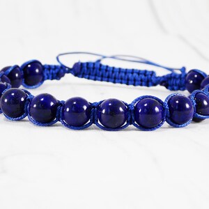 May include: A blue beaded bracelet with a knotted cord. The beads are round and dark blue, and the cord is a lighter shade of blue.
