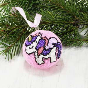 May include: Pink Christmas ornament with a beaded unicorn design. The unicorn is white with purple wings and a gold horn.