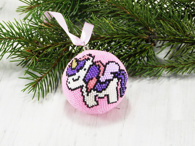 May include: Pink Christmas ornament with a beaded unicorn design. The unicorn is white with purple wings and a gold horn.