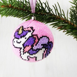 May include: Pink beaded Christmas ornament with a unicorn design. The unicorn is white with purple wings and a gold horn.