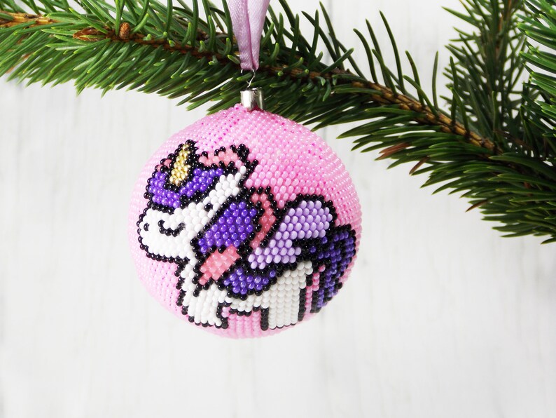 May include: Pink beaded Christmas ornament with a unicorn design. The unicorn is white with purple wings and a gold horn.