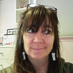 May include: A woman with brown hair and bangs wearing glasses and long, white tassel earrings. She is wearing a green shirt and looking to the right.