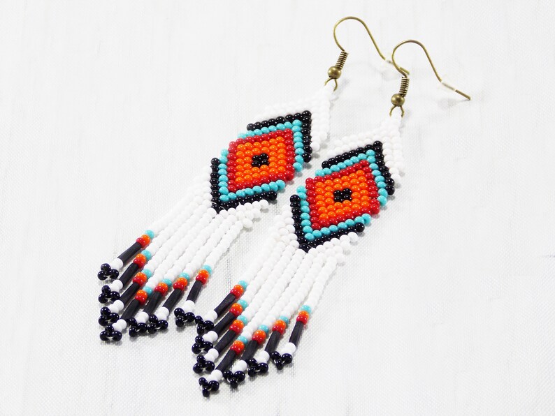 May include: White beaded earrings with a geometric design. The earrings feature a diamond-shaped pattern in orange, turquoise, and black. The fringe is white with black, red, and turquoise accents. The earrings have gold-colored hooks.
