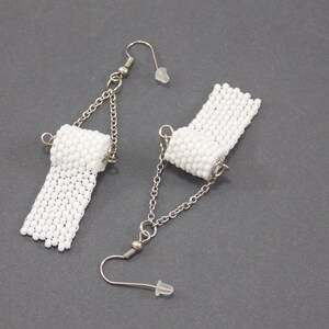 May include: A pair of white beaded earrings shaped like toilet paper rolls. The earrings are attached to silver chains with silver hooks.