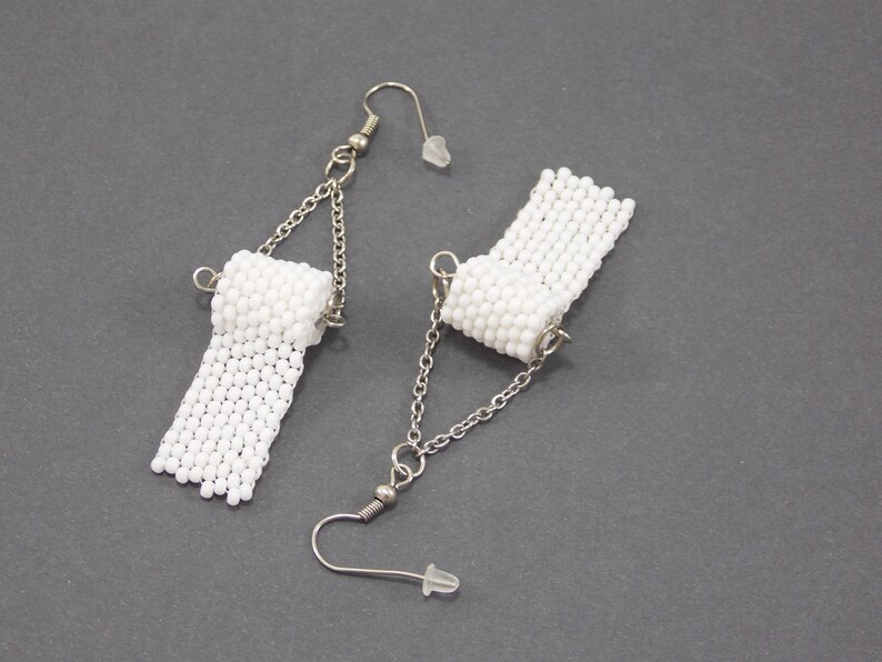 May include: A pair of white beaded earrings shaped like toilet paper rolls. The earrings are attached to silver chains with silver hooks.