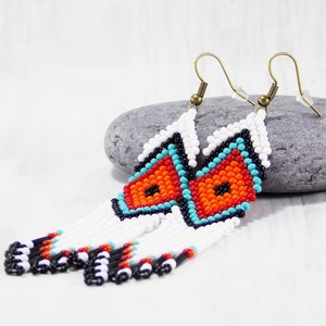 May include: Handmade beaded earrings with a geometric design. The earrings feature a white, orange, turquoise, and black pattern. The earrings have a fringe of white beads with black, red, and black accents. The earrings hang from bronze-colored ear wires.
