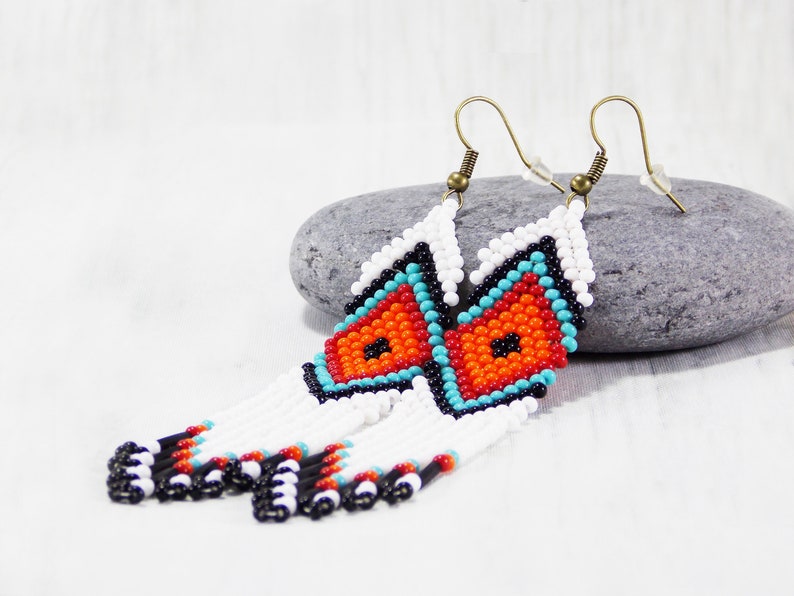 May include: Handmade beaded earrings with a geometric design. The earrings feature a white, orange, turquoise, and black pattern. The earrings have a fringe of white beads with black, red, and black accents. The earrings hang from bronze-colored ear wires.