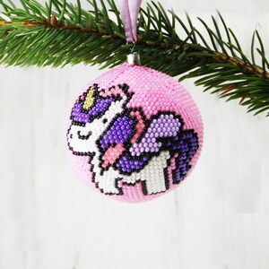 May include: Pink Christmas ornament with a beaded unicorn design. The unicorn is purple, white, and black.