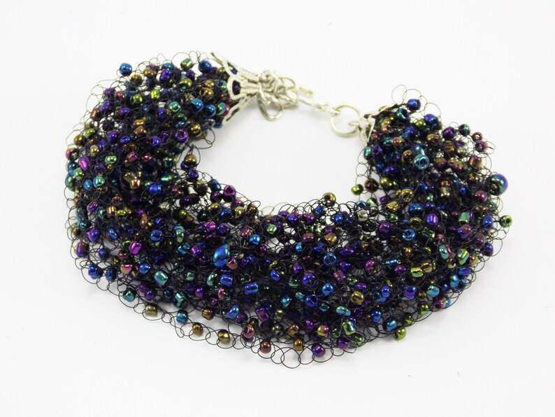 May include: A black beaded bracelet with iridescent beads in shades of blue, green, and purple. The beads are woven into a delicate net-like pattern.