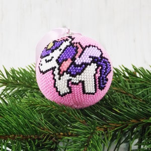 May include: Pink beaded Christmas ornament with a white unicorn design. The unicorn has a purple mane and tail.