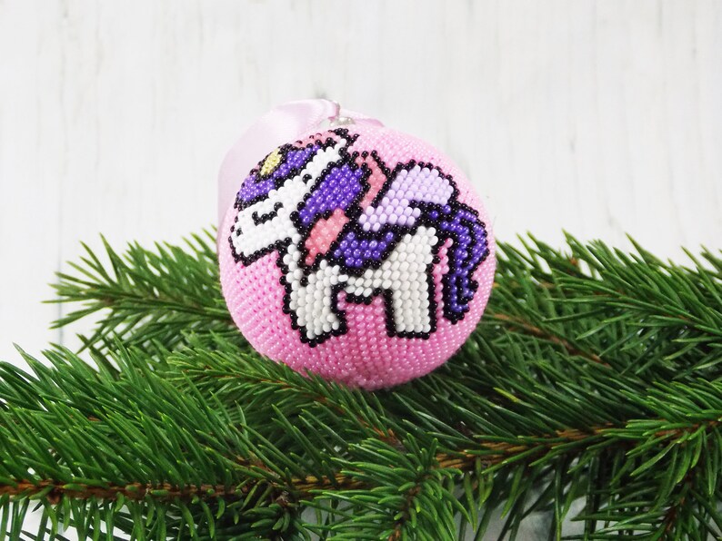 May include: Pink beaded Christmas ornament with a white unicorn design. The unicorn has a purple mane and tail.