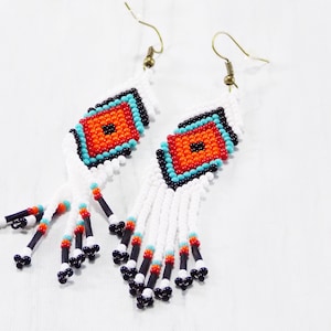 May include: Handmade beaded earrings with a geometric design. The earrings feature a central orange square surrounded by turquoise, black, and white beads. White beaded fringe hangs below, accented with black, orange, and turquoise beads.