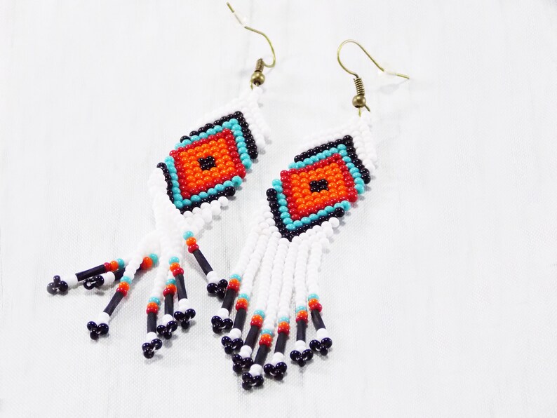 May include: Handmade beaded earrings with a geometric design. The earrings feature a central orange square surrounded by turquoise, black, and white beads. White beaded fringe hangs below, accented with black, orange, and turquoise beads.