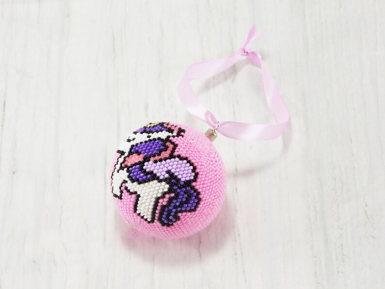 May include: Pink beaded Christmas ornament with a unicorn design. The ornament has a pink ribbon attached for hanging.