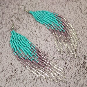 May include: A pair of long turquoise and purple beaded earrings with silver fringe. The earrings are made with small seed beads and have a teardrop shape.
