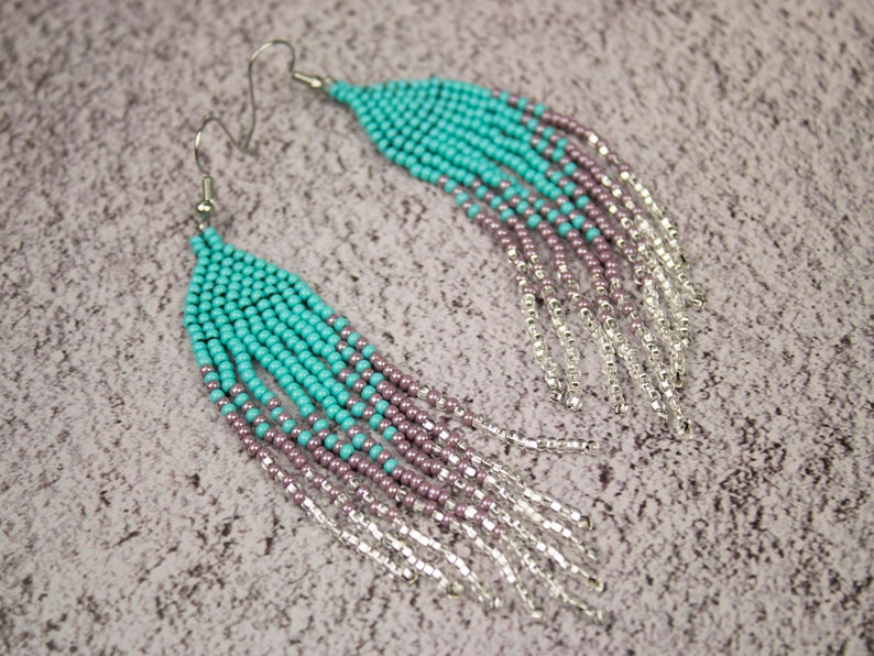 May include: A pair of long turquoise and purple beaded earrings with silver fringe. The earrings are made with small seed beads and have a teardrop shape.