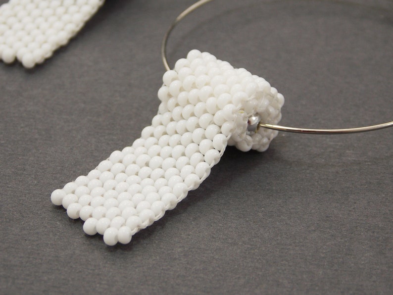 May include: A pair of white beaded earrings, each shaped like a rolled-up piece of paper. The earrings are attached to a silver hoop.