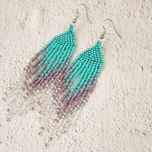 May include: A pair of long, beaded earrings with a gradient of turquoise, purple, and silver beads. The earrings are made with small, round beads and have a fringe-like design.
