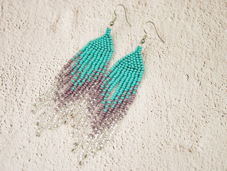 May include: A pair of long, beaded earrings with a gradient of turquoise, purple, and silver beads. The earrings are made with small, round beads and have a fringe-like design.