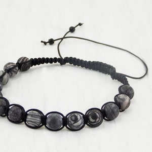 Puede incluir: Un brazalete de cord&oacute;n negro con cuentas de patr&oacute;n gris y blanco. El brazalete tiene un cierre de cord&oacute;n ajustable.