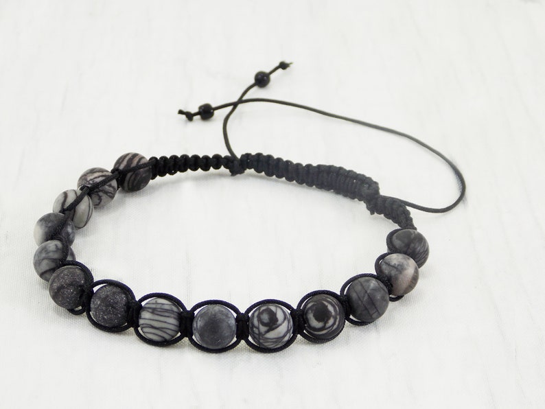 Puede incluir: Un brazalete de cord&oacute;n negro con cuentas de patr&oacute;n gris y blanco. El brazalete tiene un cierre de cord&oacute;n ajustable.