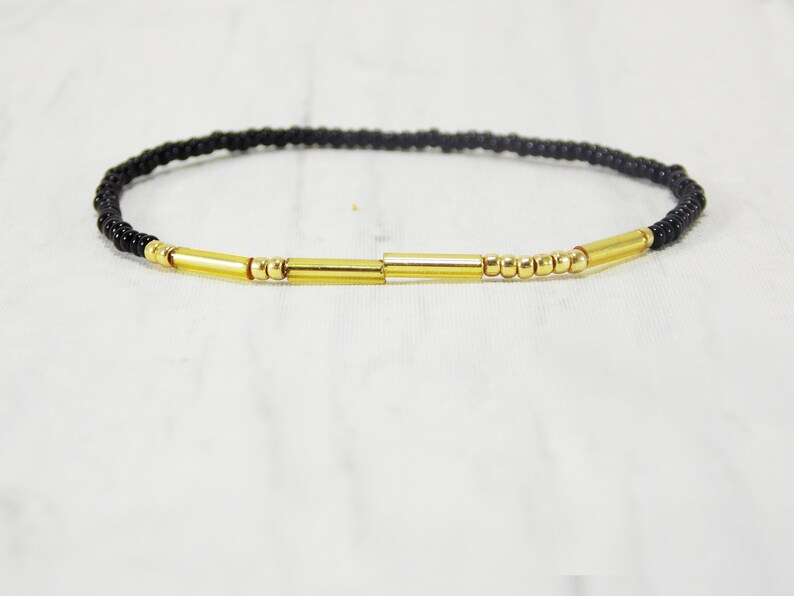 May include: A black beaded bracelet with gold accents. The bracelet has a simple design with small black beads and three gold bars.