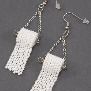 May include: A pair of white beaded earrings shaped like toilet paper rolls. The earrings are made with small white beads and have silver chains and hooks.