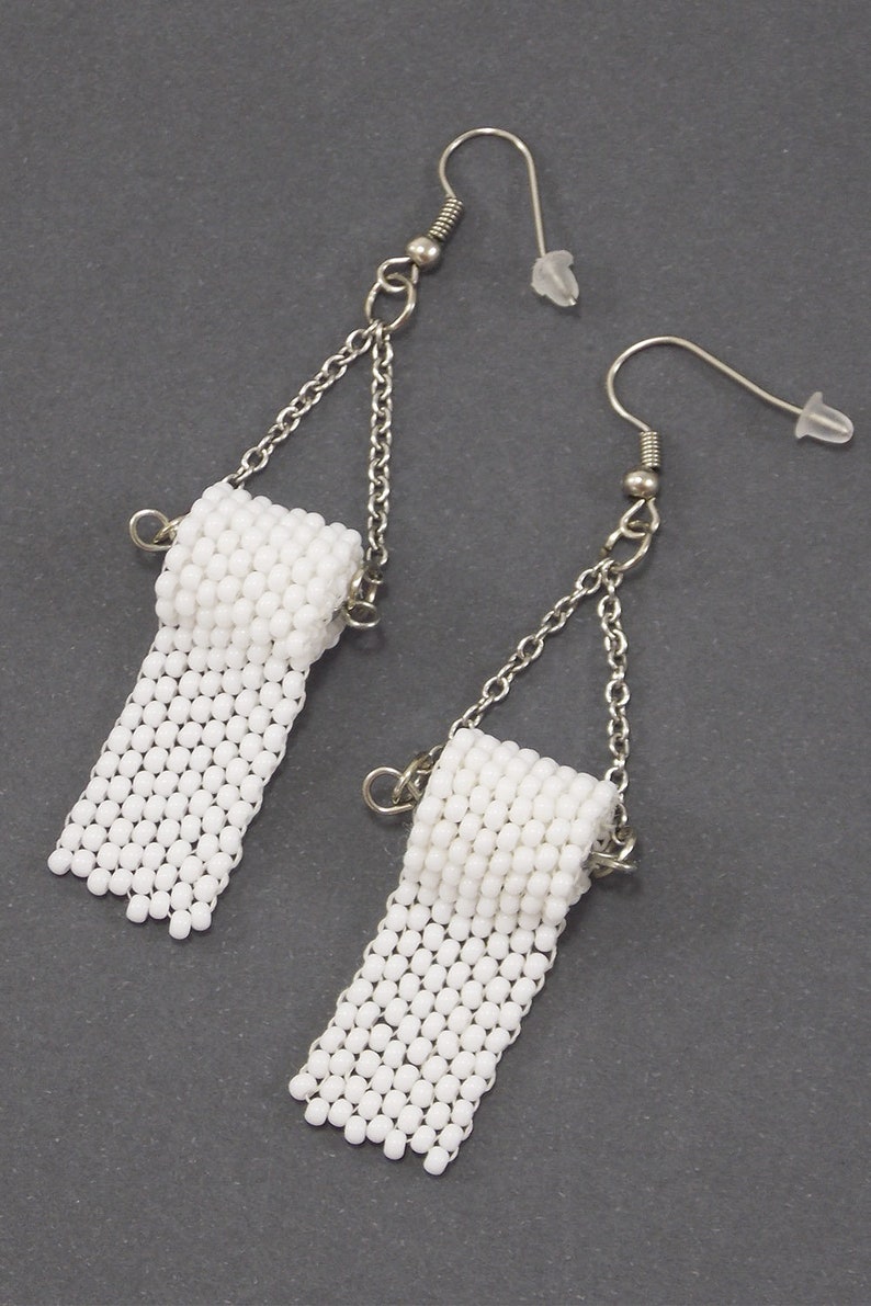 May include: A pair of white beaded earrings shaped like toilet paper rolls. The earrings are made with small white beads and have silver chains and hooks.