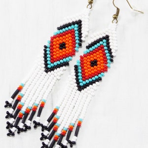 May include: White beaded earrings with a diamond-shaped design in the center. The diamond features orange, turquoise, and black beads. The earrings have white fringe with black, turquoise, and orange accents. The earrings hang from gold-colored hooks.