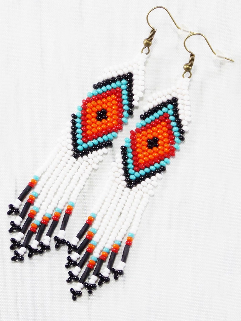 May include: White beaded earrings with a diamond-shaped design in the center. The diamond features orange, turquoise, and black beads. The earrings have white fringe with black, turquoise, and orange accents. The earrings hang from gold-colored hooks.