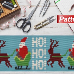 May include: A beaded pattern for a Christmas ornament featuring Santa Claus riding in a sleigh pulled by reindeer. The pattern includes the words "HO! HO! HO!" in white beads on a blue background.