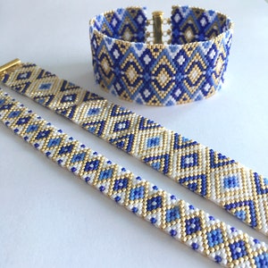 May include: Two beaded bracelets with a geometric pattern in shades of blue, white, and gold. The larger bracelet is a cuff style, while the smaller bracelet is a thinner, more delicate style.
