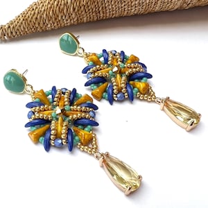 以下が含まれることがあります： 青い、黄色い、緑色のビーズで幾何学模様が施された、ゴールドトーンのイヤリングのペア。各イヤリングには、下から垂れ下がった涙滴型のクリアクリスタルが付いています。