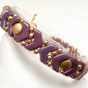May include: A purple and gold beaded bracelet with a chevron pattern. The bracelet is made with small, faceted beads and is strung on a clear elastic cord.