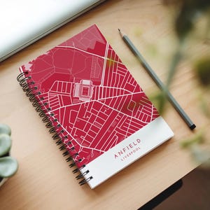 May include: A spiral-bound notebook with a red and white map of Anfield, Liverpool. The notebook has a white band at the bottom with the text "ANFIELD LIVERPOOL". A grey pencil lies beside the notebook on a light wood surface.