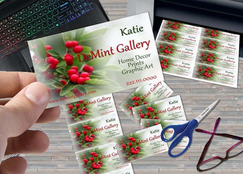 May include: A stack of business cards with a red berry and green leaf design. The text on the cards reads "DeMint Gallery", "Home Decor Prints Graphic Art", "Katie", and "222.111.0000".
