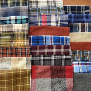 May include: A collection of folded fabric swatches featuring various patterns and colours. The assortment includes multiple plaid designs in shades of blue, green, red, brown, yellow, black, and white, alongside solid-coloured pieces in rust and beige.